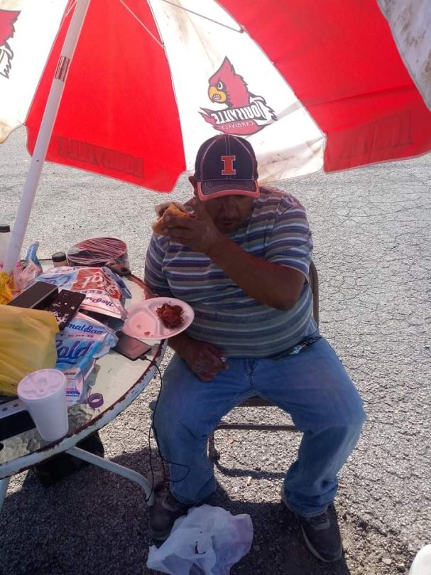 Is you hungry? He was! Man sitting under a large umbrella, eating food with a soda on a table nearby.